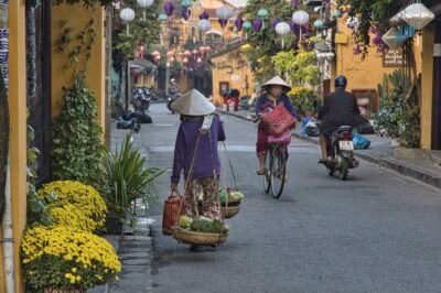 dans la rue au Vietnam une femme en vélo s'approche, une femme portant des légumes dans des paniers s'éloigne